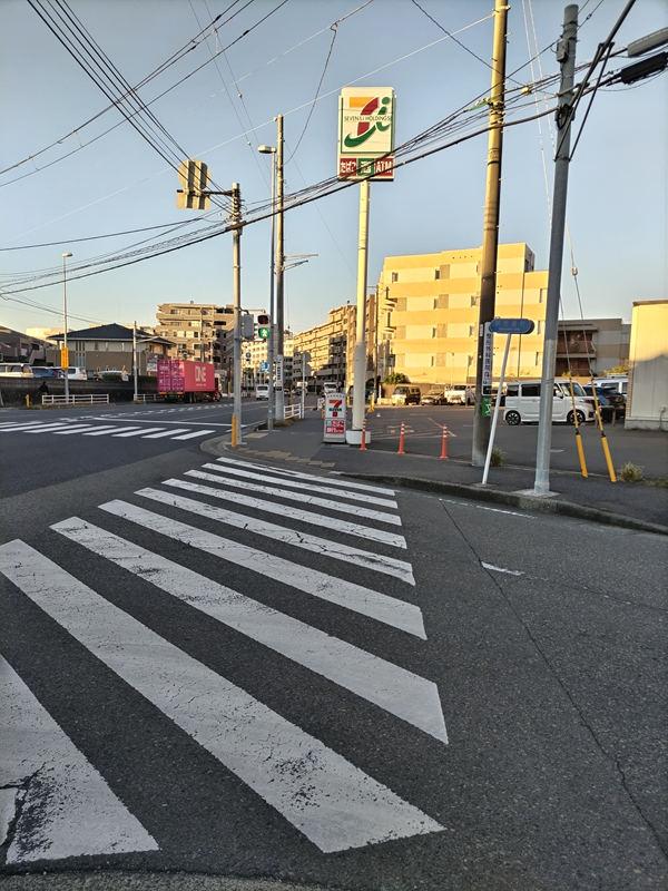 元ニート引籠りが横断歩道の先にある朝日で照らされた早朝のセブンイレブンの看板を見ている様子